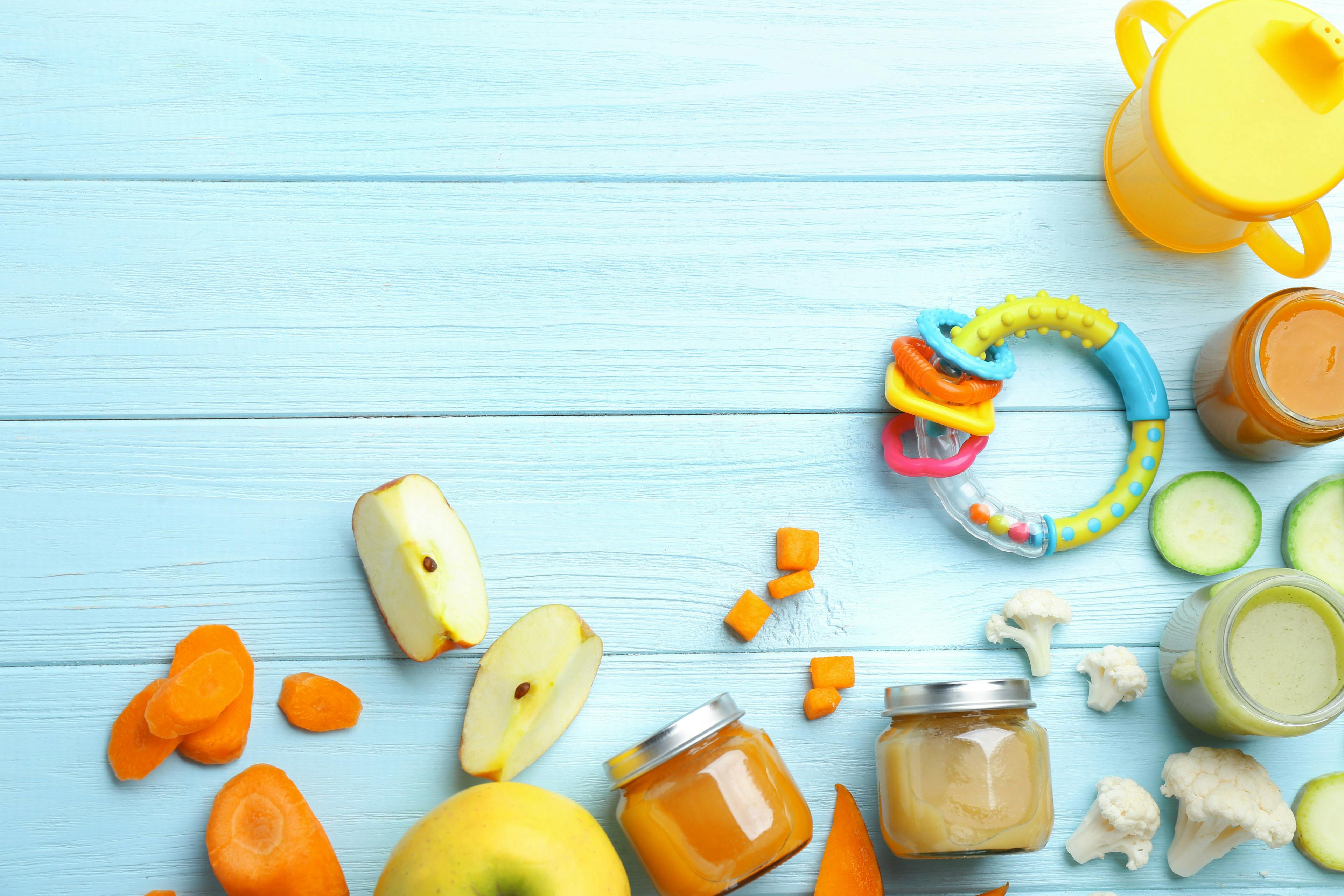 Baby food in jars, fruits, and a rattle on a light-blue table.
