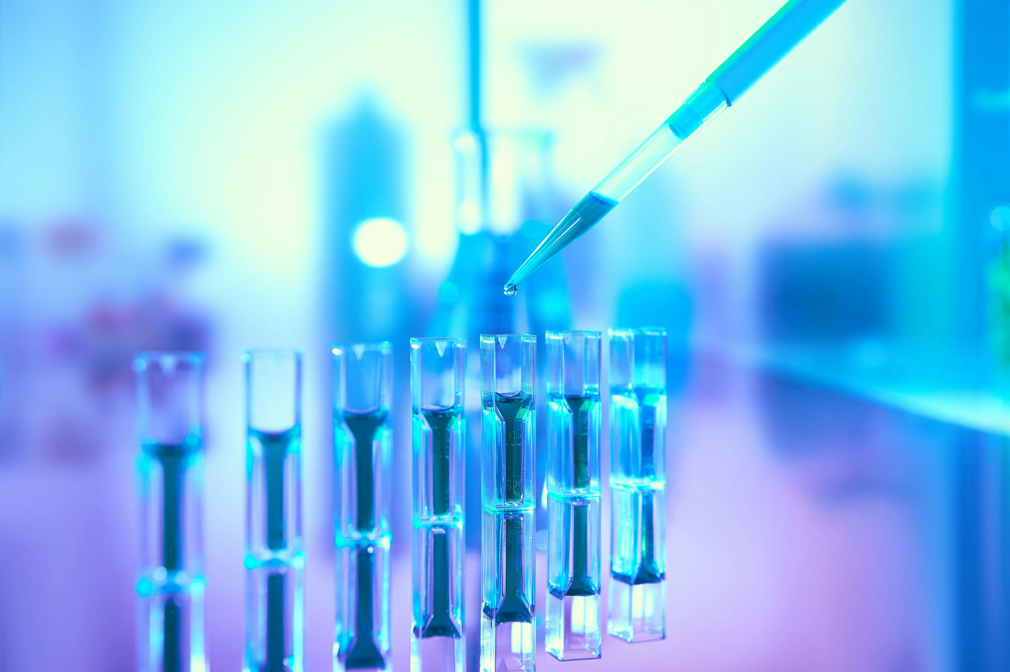 Scientific background in vibrant neon colors, purple, blue and turquoise. Pharma, biotech, protein analysis. Spectrophotometer quvettes on a reflective surface, copy-space., focus on pipette tip. | Image Credit: © tilialucida - stock.adobe.com 