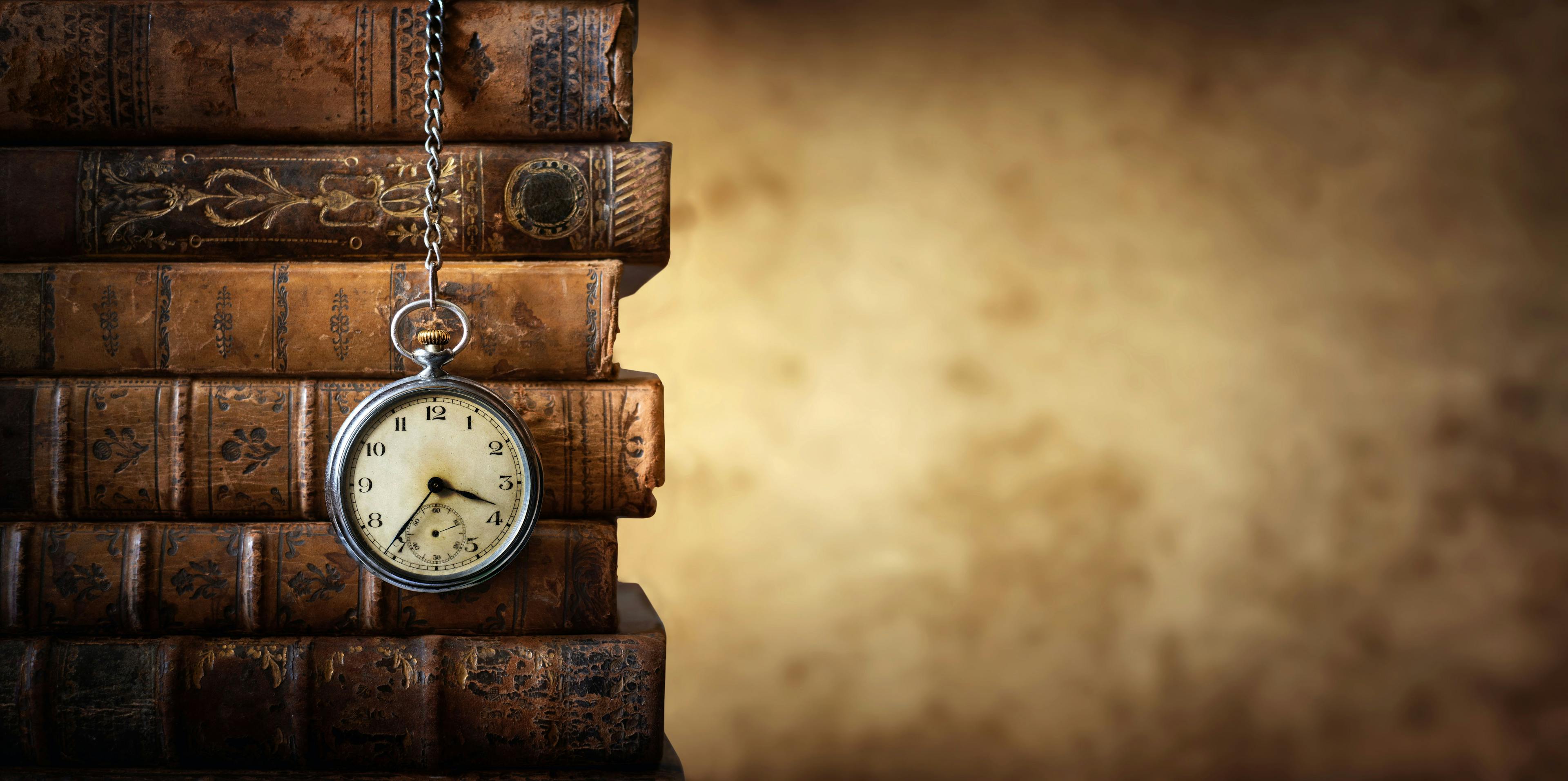 Vintage clock hanging on a chain on the background of old books. Old watch as a symbol of passing time. Concept on the theme of history, nostalgia, old age. Retro style | Image Credit: © Tryfonov - stock.adobe.com 